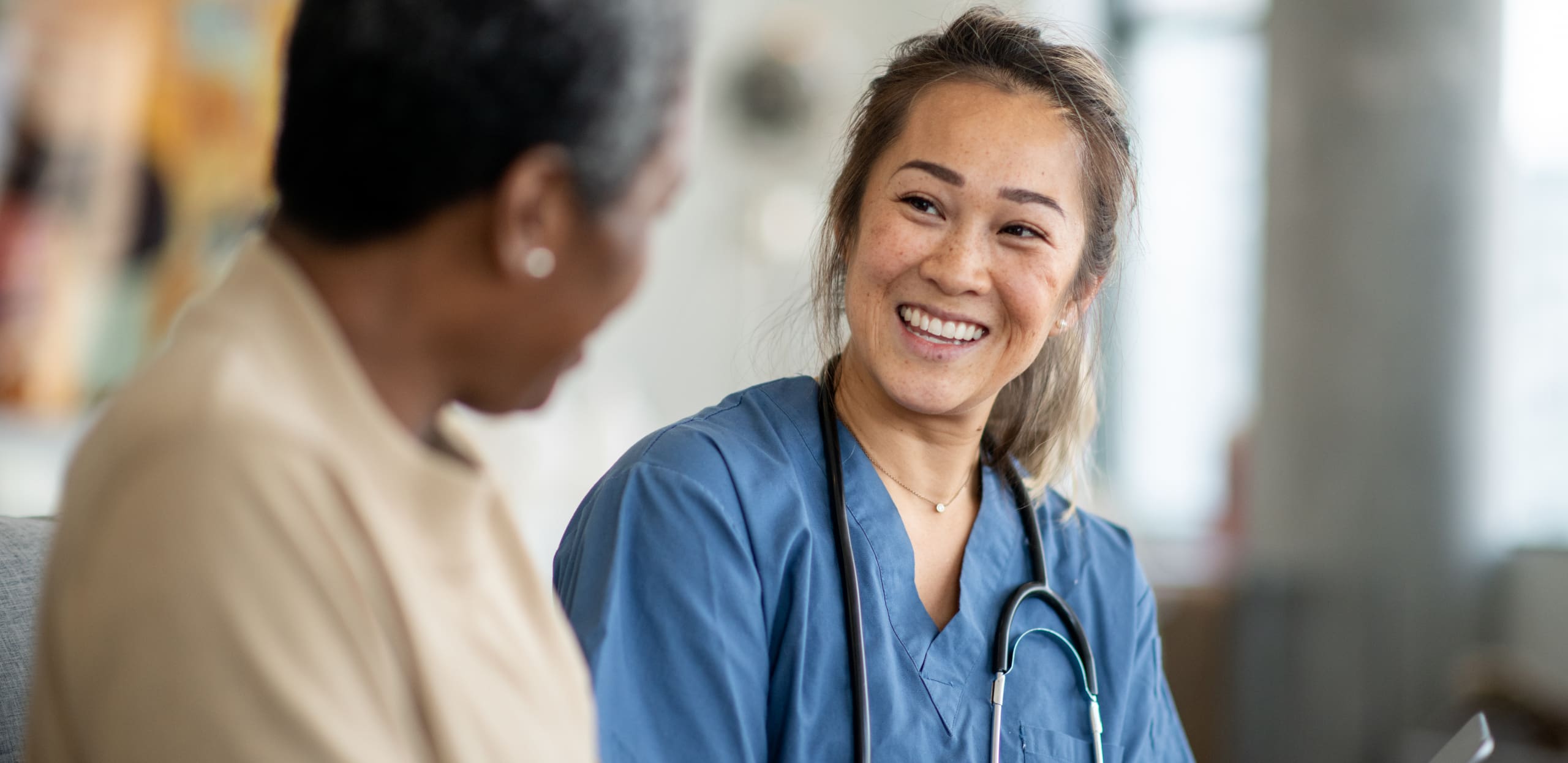 A young female healthcare worker with a stethoscope around her neck smiling warmly while talking to an older woman, both sitting indoors in a caring environment.