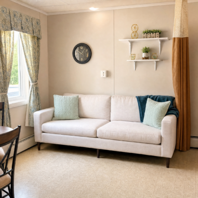 A white sofa with two light green pillows and one dark blue pillow is placed against a beige wall in a living room. To the left, there is a window with floral curtains, and a black wall clock hangs above the sofa. On the right, two white shelves hold decorative items, including a small clock, a plant, and a sign that says 