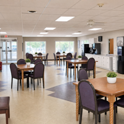 A spacious dining area with multiple wooden tables and purple upholstered chairs, large windows letting in natural light, a vending machine, and a small potted plant on one table.