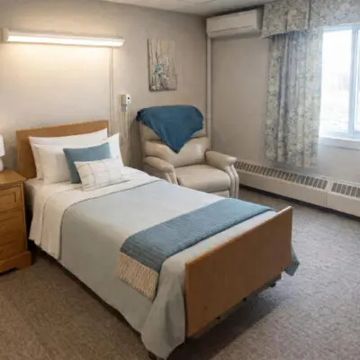 A bedroom with a bed covered in white and blue linens, a wooden nightstand, a cream armchair with a blue blanket, a window with floral curtains, and an air conditioning unit mounted on the wall.