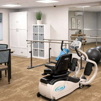 A rehabilitation room with a seated exercise machine, a white cabinet, a cubby storage unit with small drawers, a potted plant on top, and a mirror on the wall.