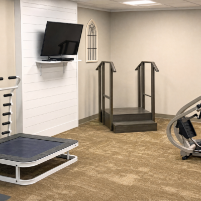 A small indoor area with a raised platform and wooden stairs with handrails, a wall-mounted TV, a therapy trampoline, and a piece of exercise equipment on the right.