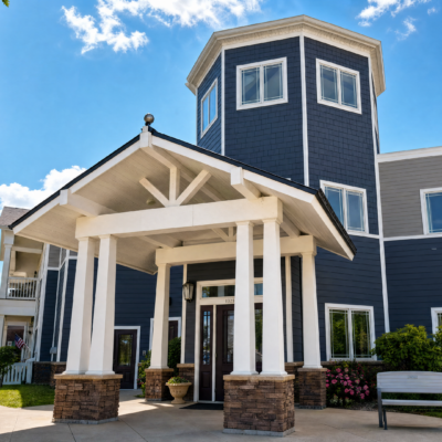 The entrance of a multi-story building with a blue exterior, white trim, and a covered porch supported by white columns. The building has a tower-like section with multiple windows, and the sky is partly cloudy.