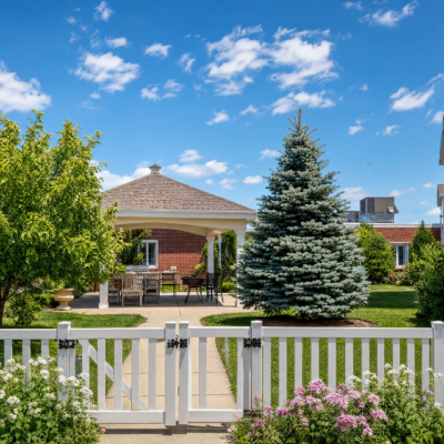 A well-maintained garden with a white picket fence, a brick gazebo with a shingled roof, lush green trees, a large blue spruce, and colorful blooming flowers under a bright blue sky with scattered clouds.