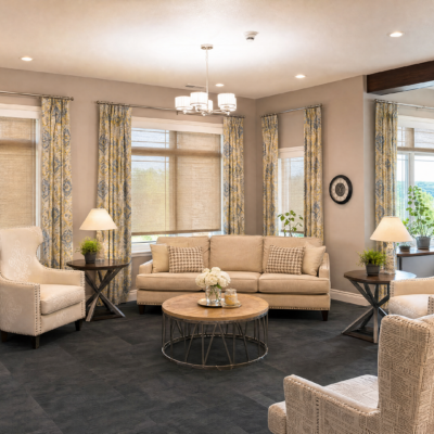 A bright, welcoming living room with a beige sofa, two matching armchairs, and a round wooden coffee table in the center. There are two side tables with lamps and potted plants, large windows with patterned curtains, and a modern chandelier hanging from the ceiling.