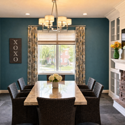 A dining room with a large rectangular marble table surrounded by eight black wicker chairs. A vase with white flowers is centered on the table. A window with floral curtains and a view of trees and a brick building outside is behind the table. A chandelier with six light fixtures hangs above the table. The room has blue walls, a white ceiling, and a white built-in cabinet with a yellow flower arrangement on the right. A wall art piece with 