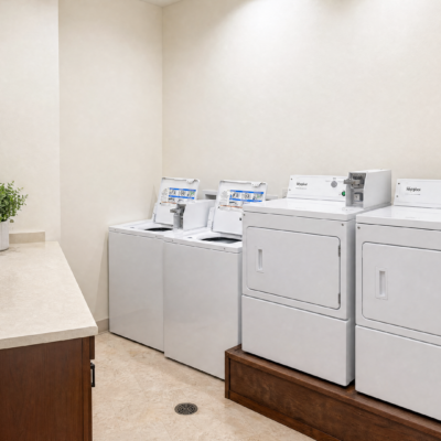 A laundry room with four white washing machines and dryers arranged side by side against a plain, light-colored wall. A small potted plant is on a countertop to the left.