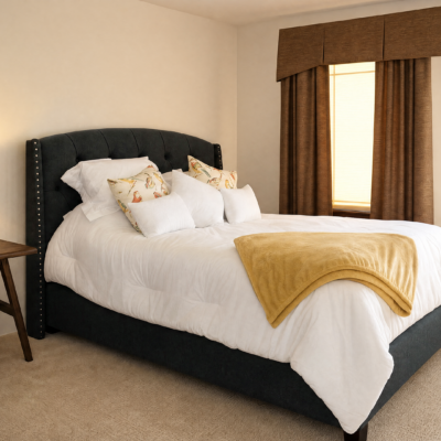 A neatly made bed with a dark upholstered headboard, white bedding, and decorative pillows. A yellow blanket is draped across the foot of the bed. There is a window with brown curtains to the right. A small wooden side table is beside the bed.