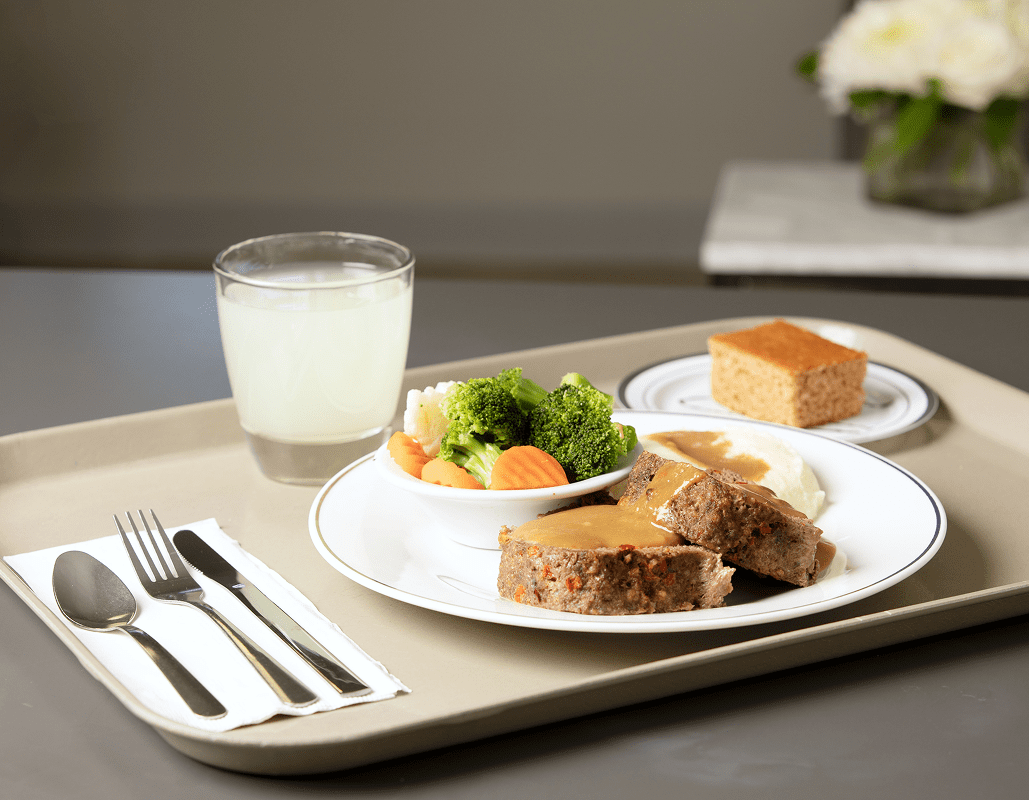 A tray with a meal consisting of meatloaf with gravy, mashed potatoes, steamed broccoli and carrots, a glass of milk, a slice of cake, and a fork and spoon on a napkin.
